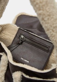 Borsa marrone in ecopelle con rivestimento interno in pelliccia sintetica color crema, dotata di una tasca con cerniera e un'etichetta del marchio argentata all'interno. Superficie esterna texturizzata.