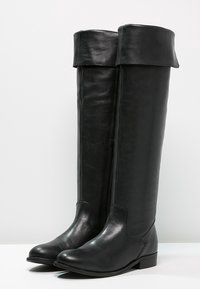 Une paire de bottes en cuir noires, hautes, avec des revers et des talons bas, se tenant côte à côte sur une surface blanche.