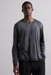 Camisa de manga longa cinza com uma textura suave, decote redondo e detalhes de costura subtil, combinada com calças pretas e joias minimalistas.