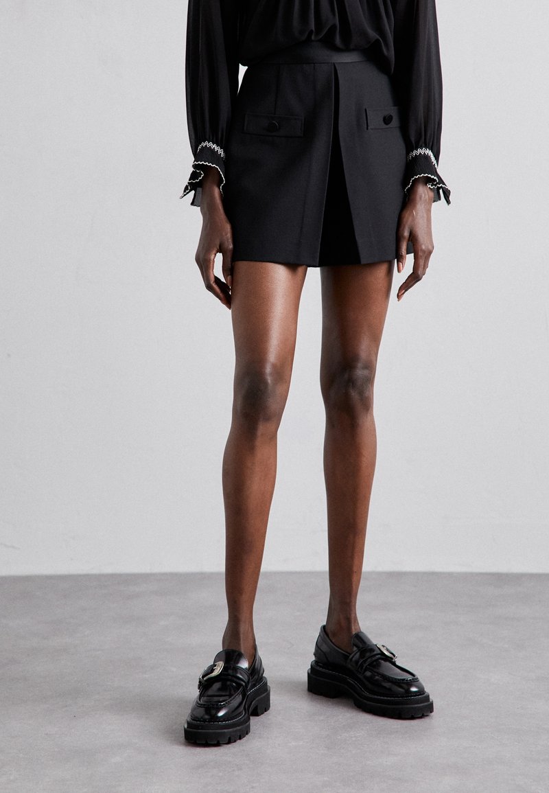 Mini-jupe noire avec pli avant, deux poches plaquées et boutons. Associée à des mocassins noirs épais. Texture lisse et design ajusté.