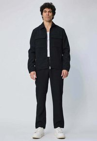 Veste noire avec deux poches poitrine, portée par-dessus un débardeur blanc, associée à un pantalon noir et des sneakers blanches. Tissu lisse, coupe décontractée.