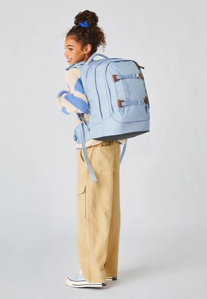 Mädchen trägt beige Hose und blau gestreiften Pullover, trägt einen großen hellblauen Rucksack, steht seitlich und lächelt.