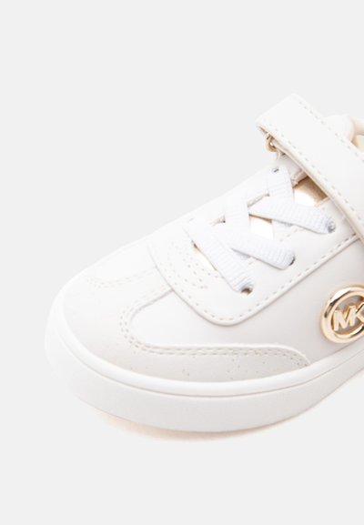 Λευκό παιδικό αθλητικό παπούτσι με κορδόνια, λουράκι Velcro και χρυσό κυκλικό λογότυπο στο πλάι, σε απλό ανοιχτό φόντο.