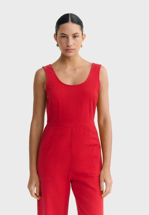 Mujer con cabello oscuro recogido en un moño, con pendientes de aro dorados y un mono rojo sin mangas, posando frente a un fondo blanco liso.