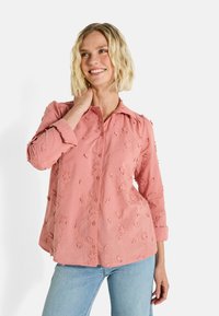 Blouse rose brodée avec un devant à boutons, col, manches trois-quarts et détails floraux, assortie avec un jean en denim clair.