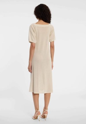 Femme aux cheveux bouclés portant une robe midi texturée beige avec des manches bouffantes et des sandales à talons hauts beiges, vue de dos sur un fond blanc.