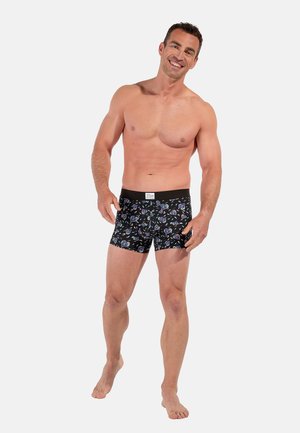 Fit Mann, der lächelt, barfuß steht und schwarze Boxershorts mit bunten abstrakten Mustern vor einem schlichten hellen Hintergrund trägt.