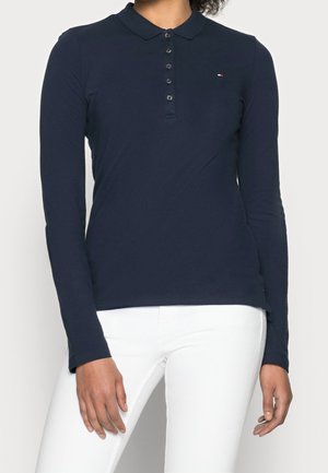 Personne portant un polo bleu marine à manches longues avec boutons et petit logo, associé à un pantalon blanc, debout devant un fond clair.