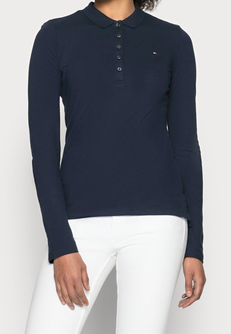 Personne portant un polo bleu marine à manches longues avec boutons et petit logo, associé à un pantalon blanc, debout devant un fond clair.