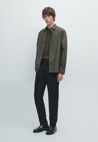 Olivově zelená košile na knoflíky přes tmavě zelenou košili, doplněná tmavými kalhotami a černými mokasínami; hladká textura a přiléhavý design.