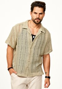 Beige gestricktes Kurzarmhemd mit einem offenen Webdesign, Kragen und Knöpfen vorne. Das Model kombinierte es mit hellen Hosen.