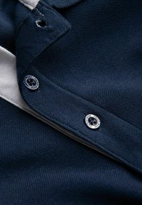 Mörkblått strukturerat tyg med en vit rand, med en metallknapp med ingraverade detaljer och en stickad krage.