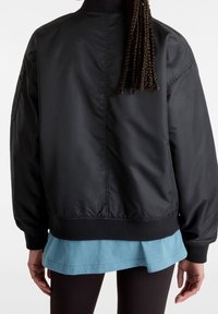 Blouson aviateur noir à fini lisse et brillant, col et poignets côtelés, avec une chemise bleue superposée en dessous.