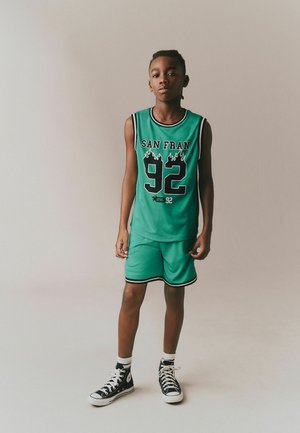 Boy standing in green sports jersey and shorts with "San Fran 92" text, wearing black high-top sneakers against plain background.