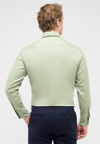 Camicia a maniche lunghe di un verde chiaro con una texture liscia, polsini abbottonati e colletto classico; abbinata a pantaloni navy con tasche posteriori.