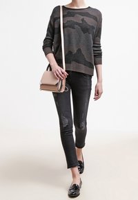 Strickpullover mit einem dunklen Tarnmuster, kombiniert mit schwarzen, distressed Skinny-Jeans. Eine braune Umhängetasche und schwarze Loafer runden das Outfit ab.