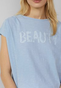 Lichtblauw T-shirt met korte mouwen van een licht gestructureerde stof; met het woord "SCHOONHEID" in een zachte, lichte print op de voorkant.