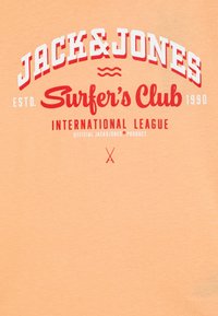 Persiko spalvos marškinėliai, pagaminti iš minkštos medvilnės. Su ryškiu raudonu ir baltu ekrano spausdintu tekstu "JACK&JONES Surfer's Club." Paprastas dizainas, be raštų.