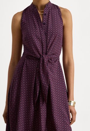 Ærmeløs lilla kjole med rødt geometrisk mønster, V-hals, knudedetalje ved taljen, båret med guldbøjle og delikat halskæde.