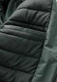 Chaqueta acolchada de tela verde oscuro, con secciones acolchadas, forro suave y un bolsillo con cremallera en el interior para un almacenamiento seguro.