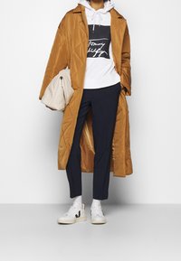 Abrigo largo, acolchado y marrón sobre una sudadera blanca con un logo negro, combinado con pantalones azul marino y zapatillas blancas. También se incluye un bolso texturizado beige.