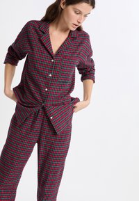 Mujer vistiendo pijamas de cuadros rojos y verdes con botones y mangas arremangadas, ajustando el dobladillo inferior contra un fondo blanco.