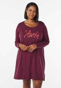 Donna che indossa un vestito rosso bordeaux a maniche lunghe con motivi di "amore" e cuori corallo sul petto, in piedi davanti a uno sfondo bianco.