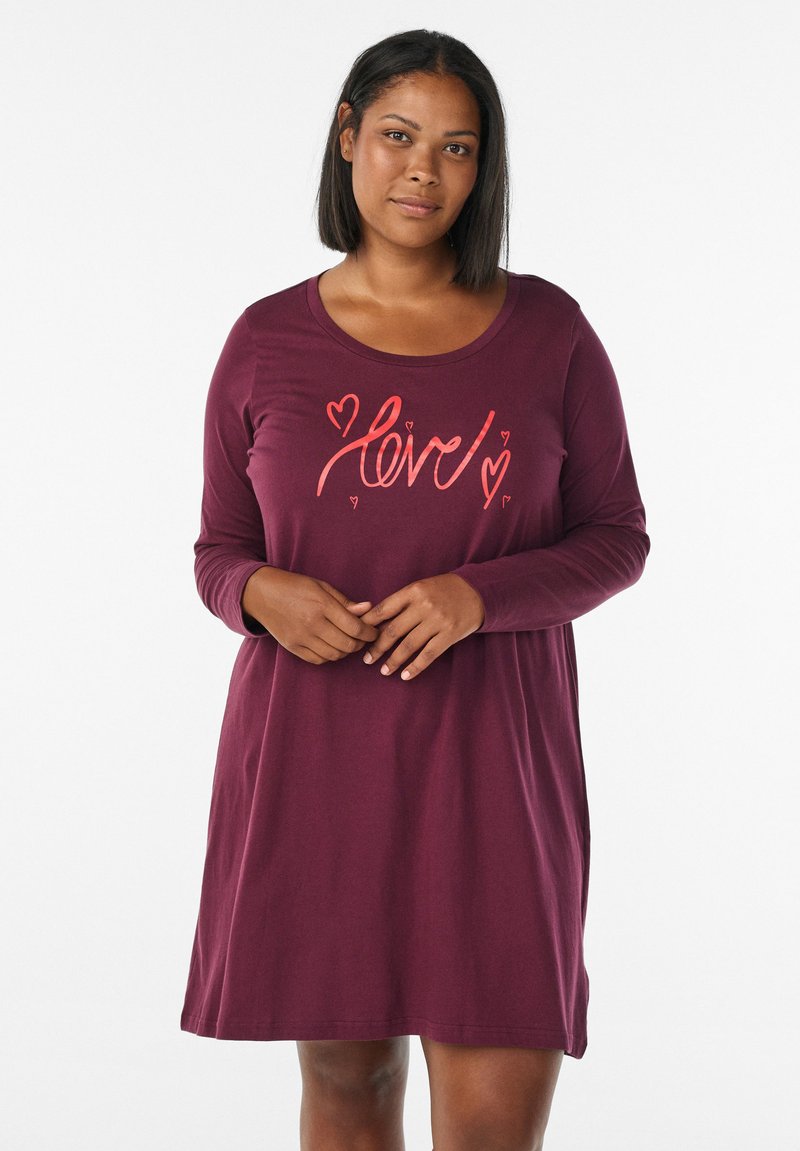 Donna che indossa un vestito rosso bordeaux a maniche lunghe con motivi di "amore" e cuori corallo sul petto, in piedi davanti a uno sfondo bianco.