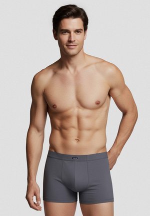 Sportlicher Mann mit kurzen braunen Haaren, der graue Boxershorts trägt, steht mit einer Hand hinter dem Rücken vor einem einfarbigen hellen Hintergrund.
