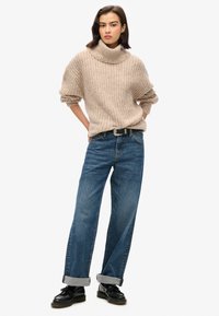 Suéter de punto beige con cuello alto y corte holgado, combinado con jeans de denim azul de pierna recta, remangados en los dobladillos, y zapatos negros de cordones.