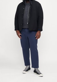 Czarna ocieplana kurtka na guziki, szary sweter o fakturze, granatowe spodnie chino oraz czarne sneakersy za kostkę z białymi akcentami.