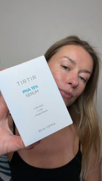 Mujer de piel clara y cabello rubio sostiene una caja de suero TIRTIR PHA 15% cerca del rostro sobre un fondo liso.