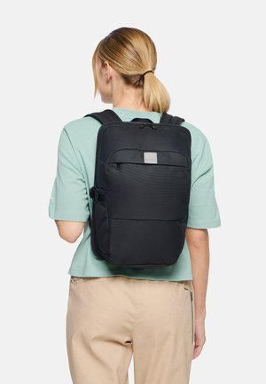 Vrouw met blond paardenstaart, gekleed in een zwart rugzak en een lichtgroene blouse, met de rug naar de camera toe tegen een effen witte achtergrond.