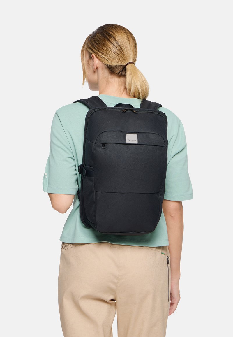 Mujer con coleta rubia, lleva una mochila negra y una camisa verde claro, de espaldas sobre un fondo blanco liso.