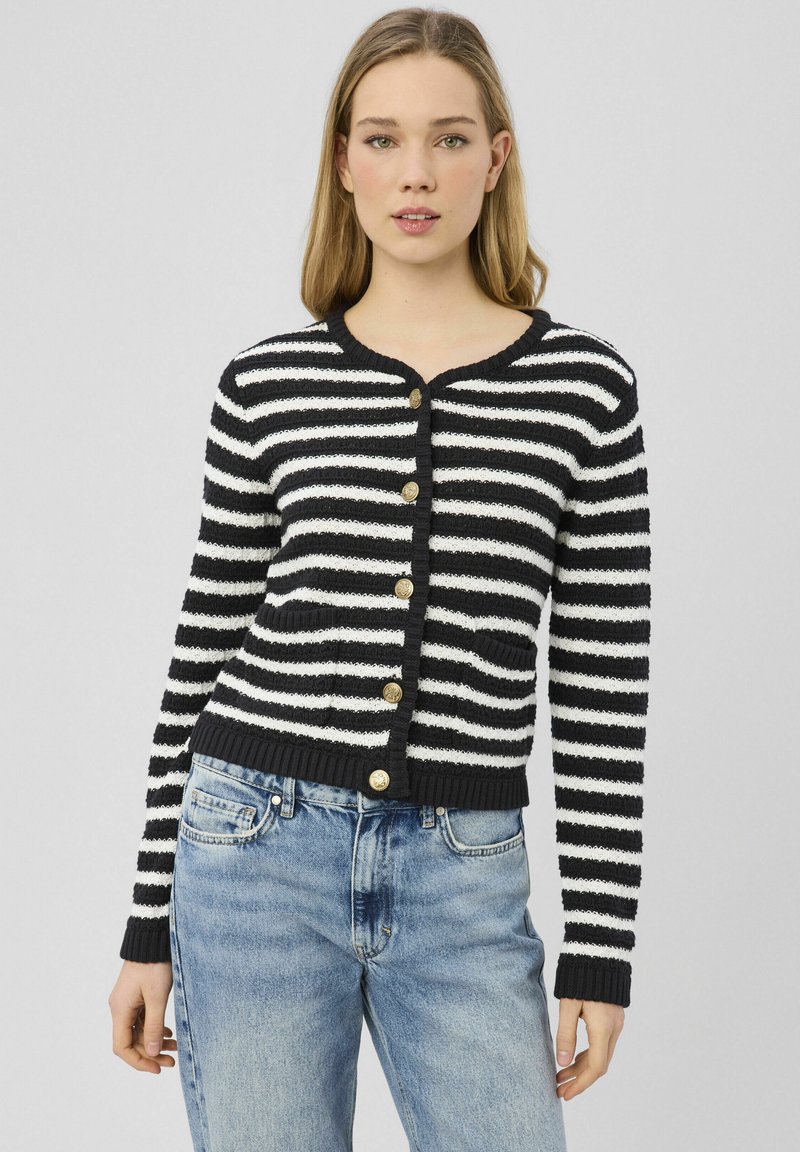 Jeune femme portant un cardigan noir et blanc rayé avec des boutons dorés et un jean en denim bleu clair, debout devant un fond uni.