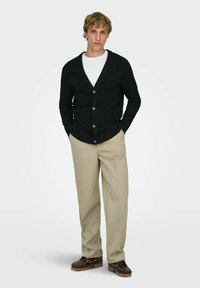 Young man standing with hands in pockets wearing black buttoned cardigan, white shirt, beige trousers, and brown shoes against a plain background.