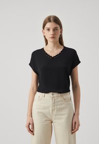 Svart kortermet topp med bølget V-hals, laget av lett stoff. Brukt sammen med kremfargede høyt midje jeans.