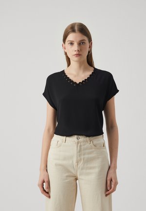 Black short-sleeve top with a scalloped V-neckline, made of lightweight fabric. Worn with cream-colored high-waisted jeans.