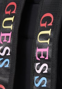 Fekete szövetpántok, amelyeken több színű "GUESS" felirat található különböző betűtípusokban. A textúra sima, enyhe fényességgel.