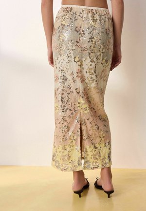 Widok z tyłu długiej spódnicy cekinowej w pastelowy kwiatowy wzór z rozcięciem pośrodku z tyłu, noszona z czarnymi sandałami na niskim obcasie.