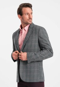 Blazer grigio a motivo houndstooth con accenti a quadri rosa, design monopetto, revers in taglio, e due tasche frontali, indossato su una camicia rosa.