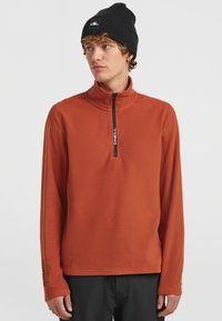 Fleece-pullover i bränd orange med halvdragkedja, med svart dragkedja och "O'Neill"-logotyp på kragen. Långa ärmar, mjuk textur.