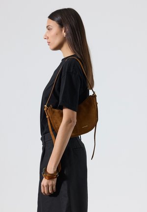 Femme portant un t-shirt noir et un pantalon, elle porte un sac à bandoulière en daim marron et porte des bracelets épais de couleur ambre à son poignet.