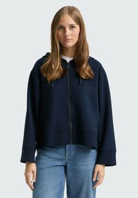 Sweat à capuche zippé marine avec une coupe décontractée, des manches larges et des cordons de serrage ajustables. Porté par-dessus un T-shirt blanc. Bas en denim décontractés.