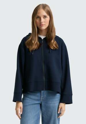 Sweat à capuche zippé marine avec une coupe décontractée, des manches larges et des cordons de serrage ajustables. Porté par-dessus un T-shirt blanc. Bas en denim décontractés.