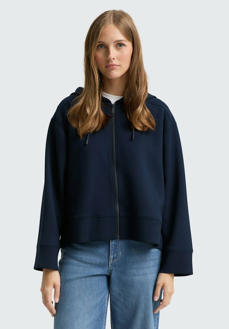 Sweat à capuche zippé marine avec une coupe décontractée, des manches larges et des cordons de serrage ajustables. Porté par-dessus un T-shirt blanc. Bas en denim décontractés.