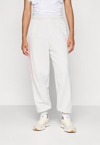 Person som har på seg hvite joggebukser med rosa striper og logo, sammen med beige og hvite sneakers, som står mot en ensfarget bakgrunn.