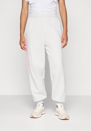 Person som har på seg hvite joggebukser med rosa striper og logo, sammen med beige og hvite sneakers, som står mot en ensfarget bakgrunn.