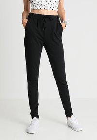 Pantaloni jogger neri con vita regolabile e tasche laterali, realizzati in un tessuto morbido, abbinati a sneakers bianche e a un crop top a pois.