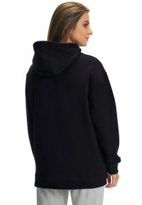 Felpa nera oversize con cappuccio regolabile, texture morbida, maniche lunghe e polsini a costine, mostrata da dietro su uno sfondo semplice.
